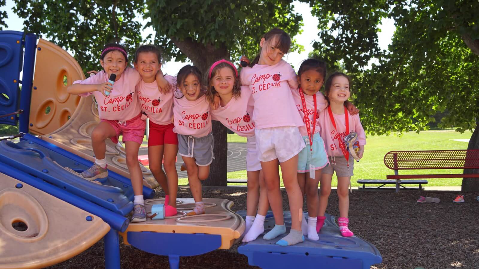 Seven young girls in matching pink Nashville summer camps shirts pose together on playground equipment, with trees and a bench visible in the background.