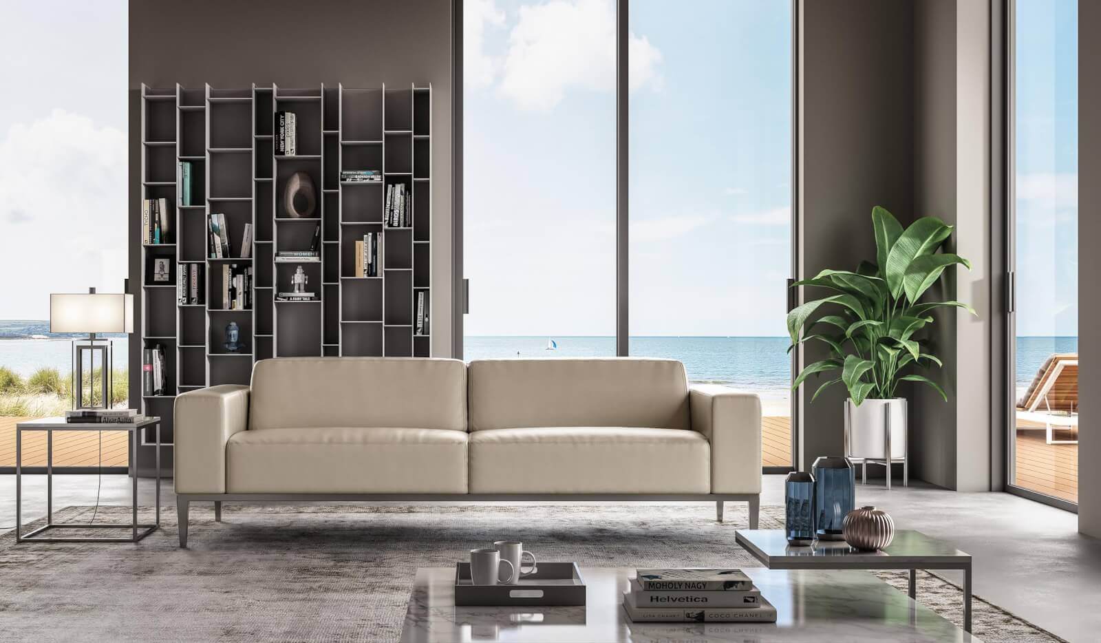 The Top Interior Designers and Architects in Birmingham - 7 Modern living room with a beige sofa, glass coffee table, and a tall plant. A metal bookshelf and lamp are on the left. Large windows reveal a seaside view with a patio—an inviting space inspired by the best interior designers and architects in Birmingham.