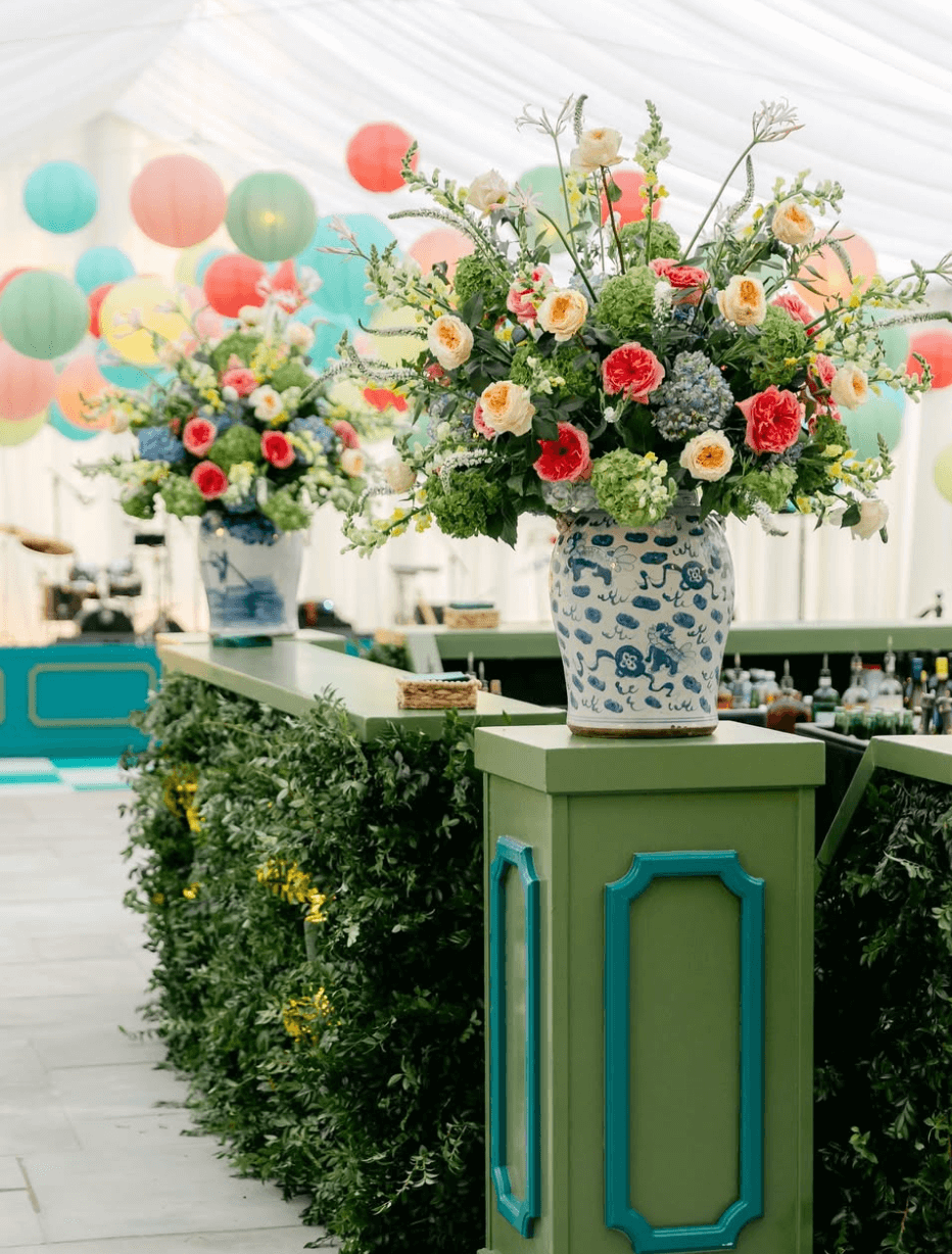 The bar counter, adorned with lush greenery and expertly arranged floral displays by Nashville florists, features painted paneling and large bouquets in blue-and-white vases. Adding a whimsical touch, vibrant paper lanterns dangle from the white tent ceiling.
