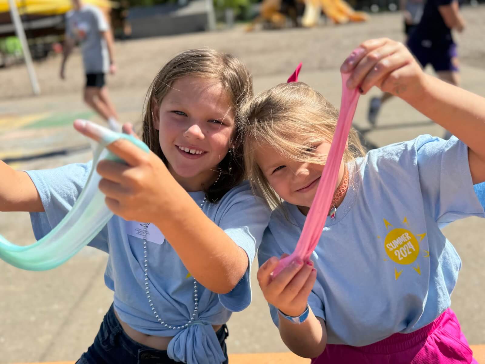 Two children wearing light blue T-shirts smile while holding up colorful slime outdoors, capturing the fun and creativity of Nashville summer camps.