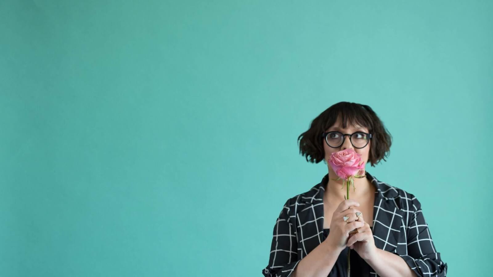 A person with glasses holds a pink rose up to their face, reminiscent of the blooms from Nashville florists. They stand against a plain teal background, wearing a black and white checkered shirt.