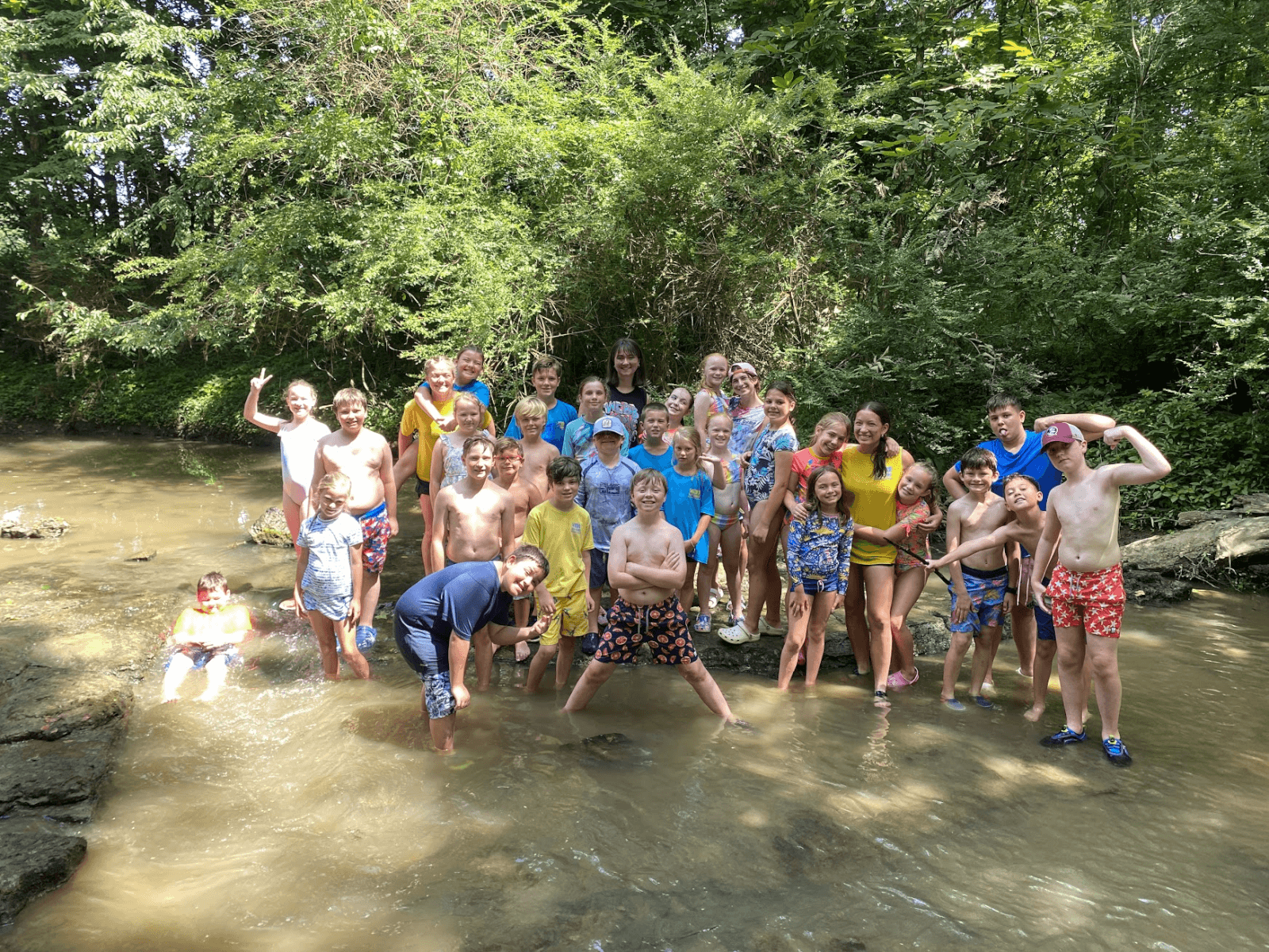 A group of children and adults from a Nashville summer camp stand and play in a shallow, wooded creek, wearing swimsuits. They are smiling and posing for the camera on a sunny day.