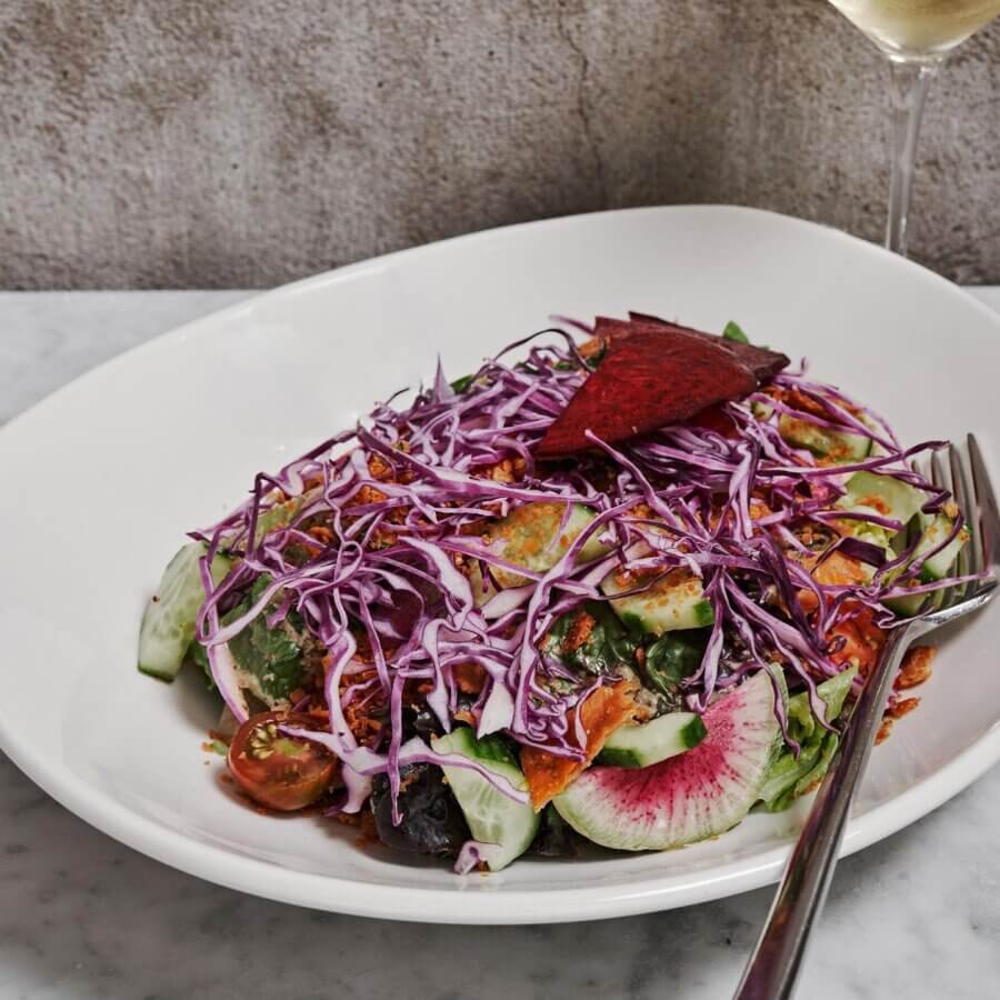 The Best Salads in Nashville: 18 Go-To Spots - 6 A white plate of mixed salad with cucumbers, cherry tomatoes, radish, shredded purple cabbage, and dressingβjust like the best salads in Nashvilleβserved next to a fork and a glass of white wine.