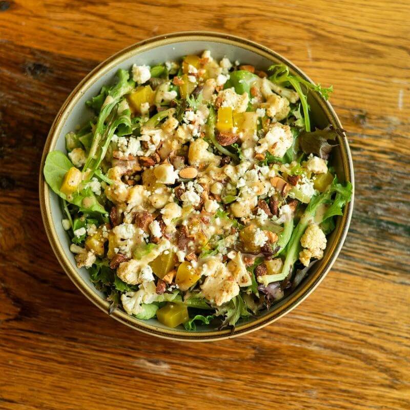 The Best Salads in Nashville: 18 Go-To Spots - 14 A bowl of mixed green salad with crumbled cheese, diced yellow peppers, nuts, and seeds on a wooden tableβjust like youβd find at one of the best salads in Nashville.