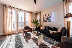Discover a modern living room on Oceola Ave, featuring a black sofa, two pink chairs, and a sleek glass coffee table under a vibrant neon guitar wall sign. Large windows draped with sheer curtains invite natural light, while a lush green plant brightens the corner.