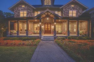 A large brick house on Stryker Place features porch lights, wooden pillars, and a landscaped front yard with flower beds and trimmed grass. The image captures the serene beauty of Brentwood, Tennessee at dusk.