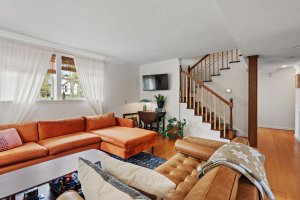 Located in a charming home on Brittany Dr, this Nashville living room features an orange sectional sofa, a wooden coffee table, and a TV on the wall. White curtains gracefully cover the window, while a houseplant adds a touch of nature near the staircase.
