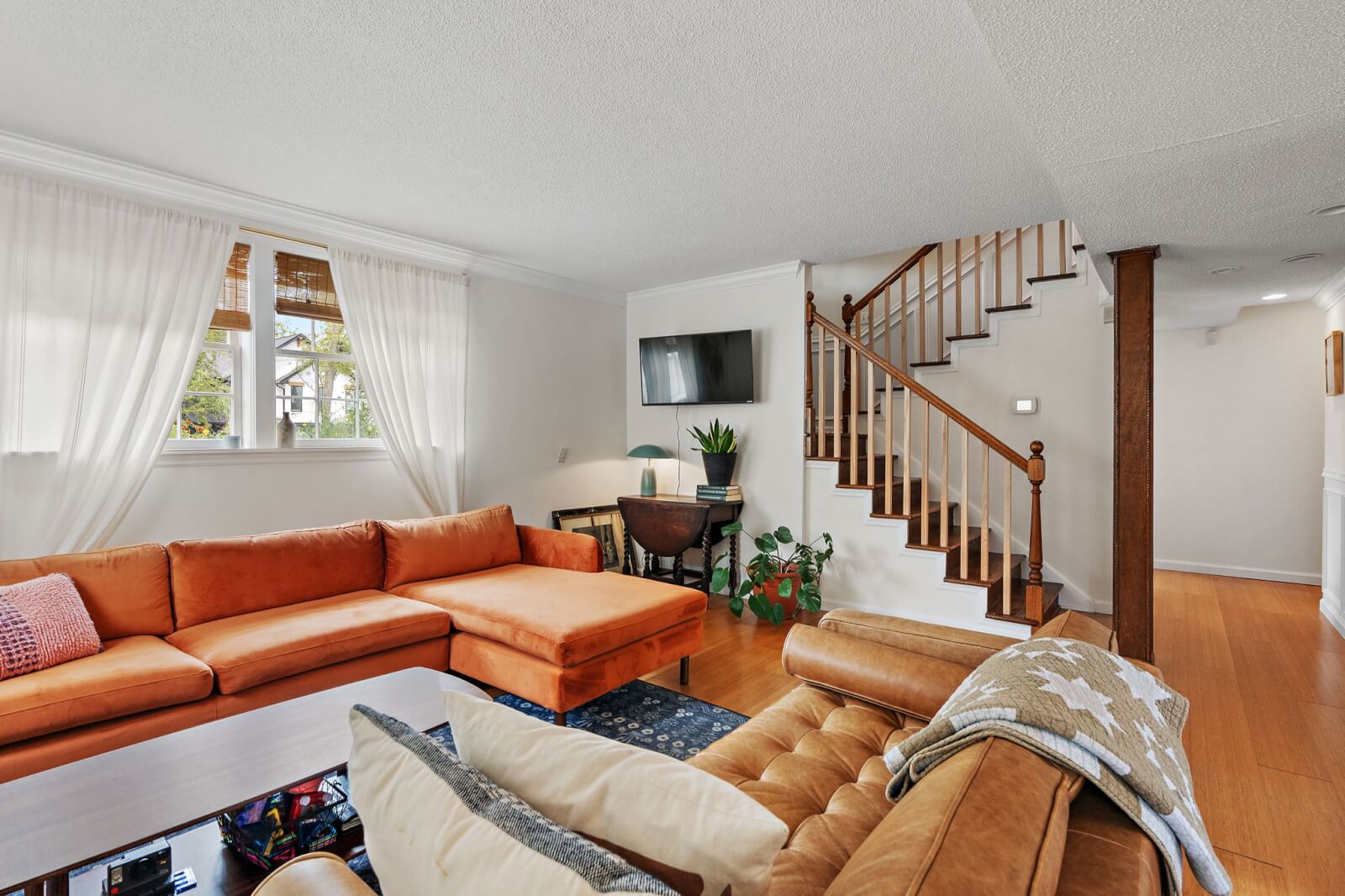 Located in a charming home on Brittany Dr, this Nashville living room features an orange sectional sofa, a wooden coffee table, and a TV on the wall. White curtains gracefully cover the window, while a houseplant adds a touch of nature near the staircase.