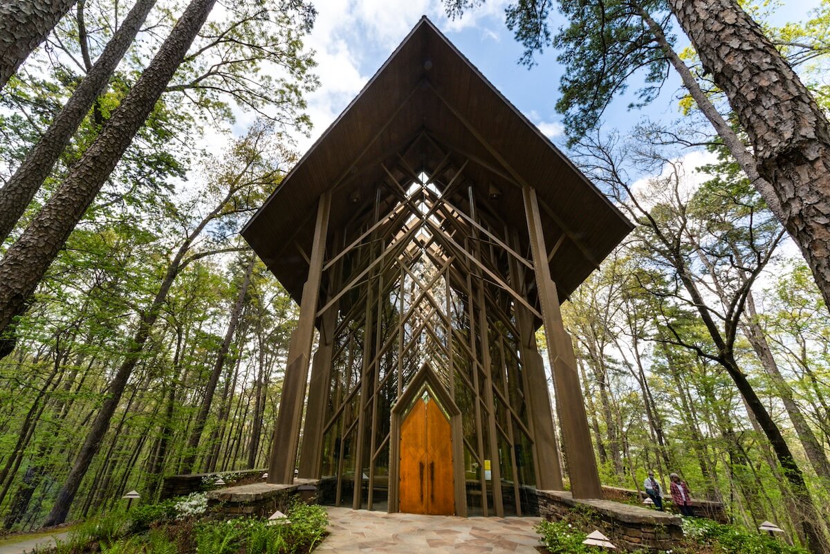 A modern chapel, reminiscent of the iconic glass chapels of Arkansas, features a tall, triangular wooden and glass structure nestled among dense trees under a cloudy sky.