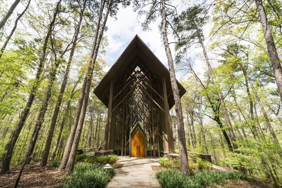 A wooden chapel with a high-peaked roof is surrounded by tall trees. Set against a backdrop of clear sky and lush greenery, a stone pathway gracefully leads to its welcoming entrance.
