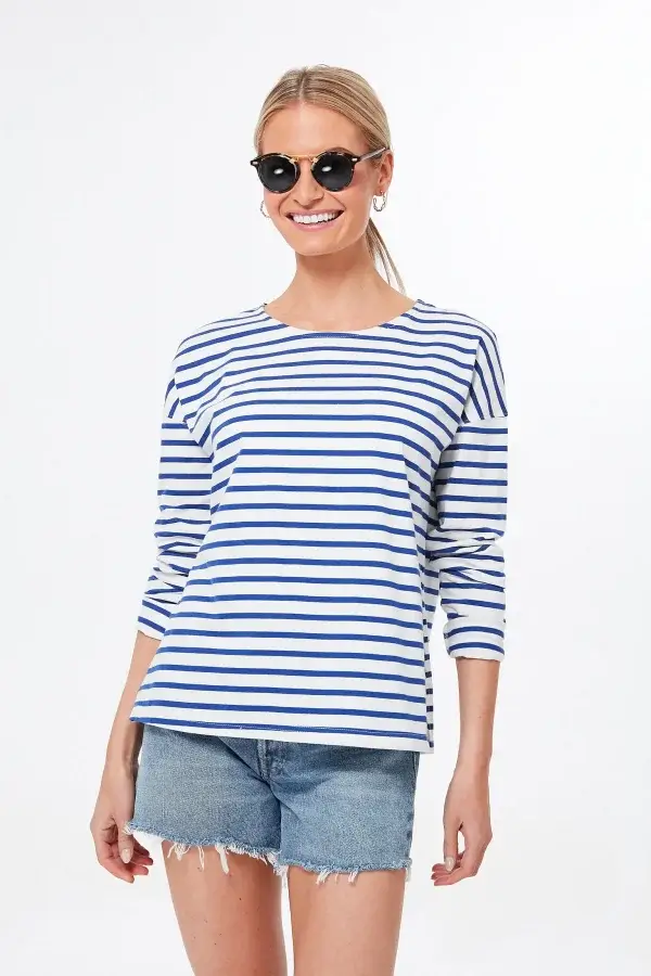 A person donning sunglasses, a blue and white striped long-sleeve shirt, and denim shorts stands confidently against a plain background, showcasing modern spin on classic pieces perfect for any spring wardrobe essentials.
