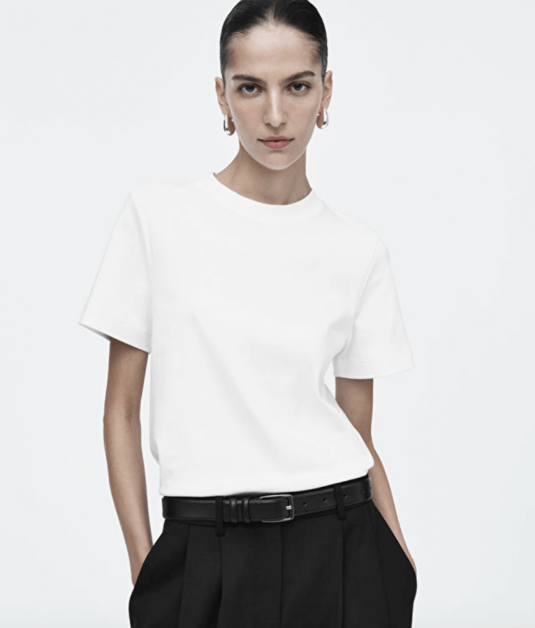 A person exudes a modern spin on classic pieces, standing with hands in pockets, wearing a white t-shirt and black pants against a plain background.