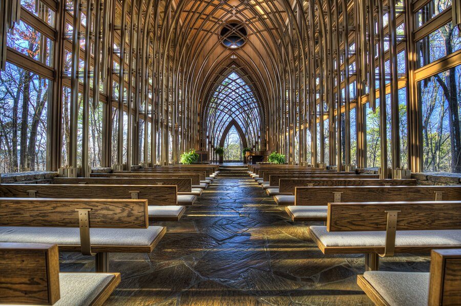 A spacious chapel with wooden pews and a high arched ceiling made of glass and wood. Large windows allow natural light in, revealing a serene forest view outside.