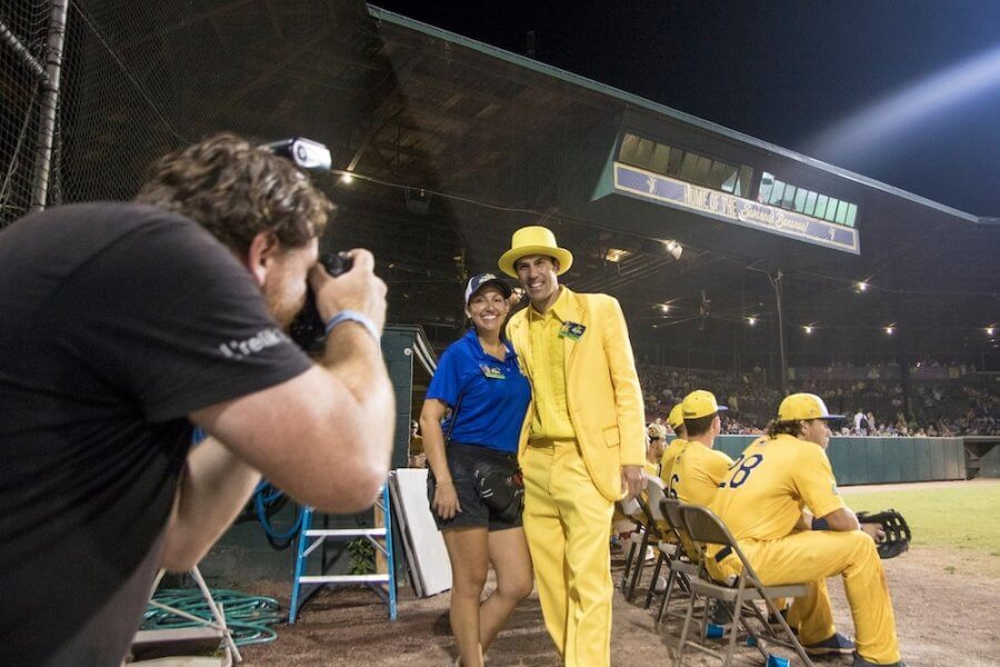 A photographer captures a woman in a blue shirt and a man in a yellow suit enjoying the Savannah Bananas baseball game, with players in matching vibrant yellow uniforms sitting nearby.