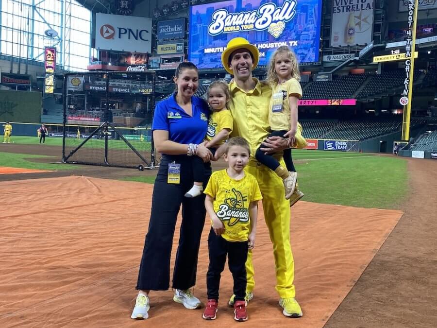 A family of five stands on a baseball field, reminiscent of a Savannah Bananas game. The father in a yellow suit holds a child, while the mother in a blue shirt is with two children in yellow shirts. Stadium seating appears behind them, capturing the lively atmosphere.