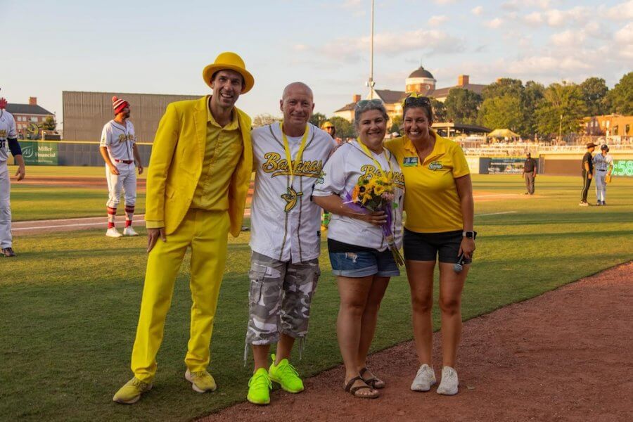 Four people strike a pose on the baseball field: one in a bright yellow suit, two in team jerseys, and another wearing a yellow shirt reminiscent of the Savannah Bananas spirit. Players and the vibrant field stretch out behind them.