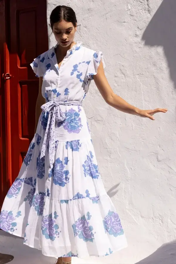 A person in a white dress adorned with blue floral patterns stands gracefully in front of a red door against a white wall, embodying the essence of a fresh spring wardrobe.