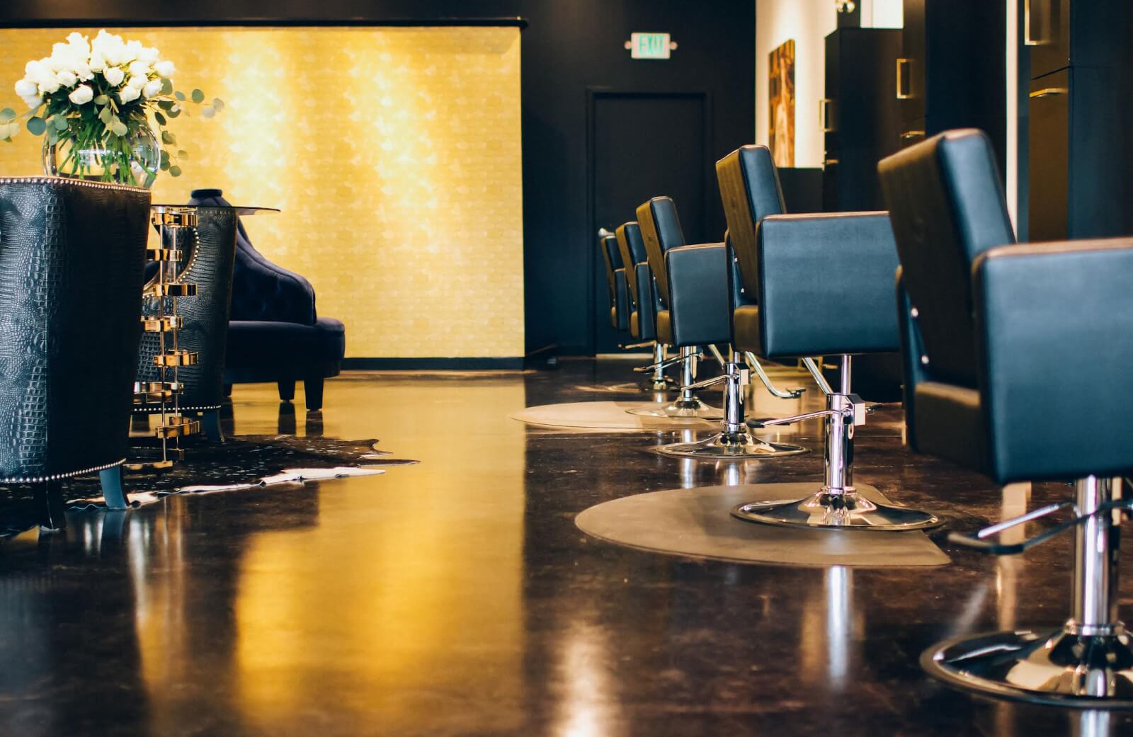 Discover one of the best hair salons in Birmingham, where modern interiors feature black styling chairs on a polished dark floor. Brightly lit walls and a vase of white flowers on the table enhance the serene ambiance, creating an ideal space for top-notch spa services.
