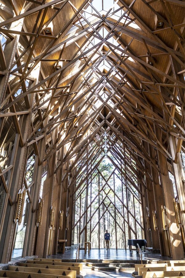 The interior of a wooden chapel in Arkansas features a high, intricate ceiling with numerous beams. Large windows let light pour in, illuminating two people standing inside.