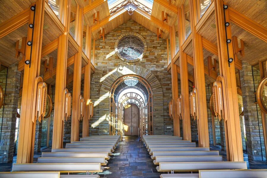 The interior of a chapel with wooden beams and stone walls echoes the serene beauty found in the glass chapels of Arkansas, as sunlight streams through arched windows, casting gentle light on the empty benches below.