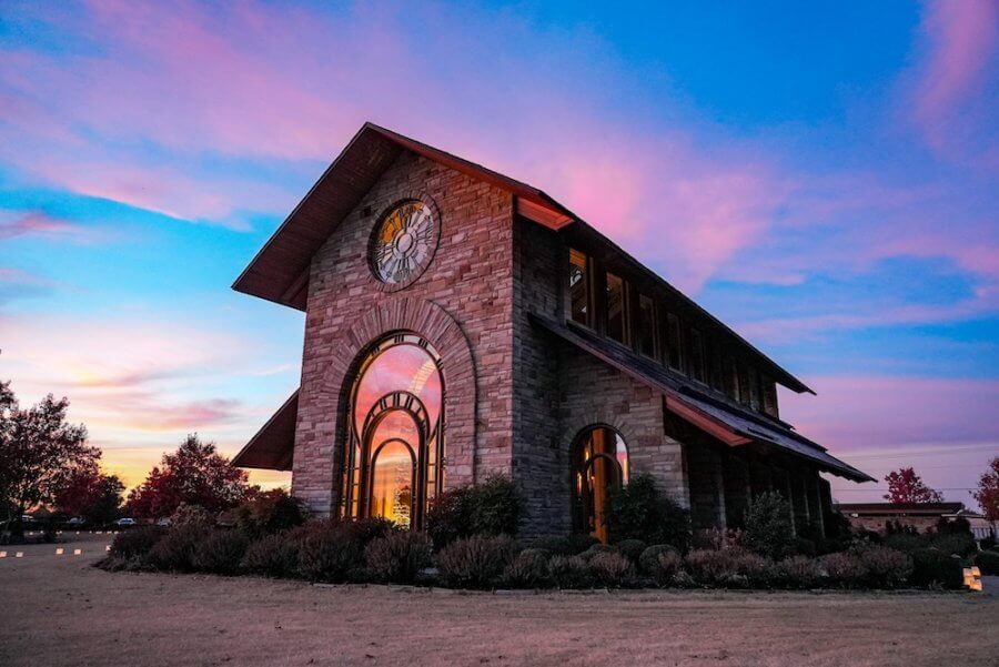 A stone building with large arched windows reminiscent of the glass chapels of Arkansas stands proudly, its clock keeping time as it is surrounded by shrubs, all set against a vibrant sunset sky.