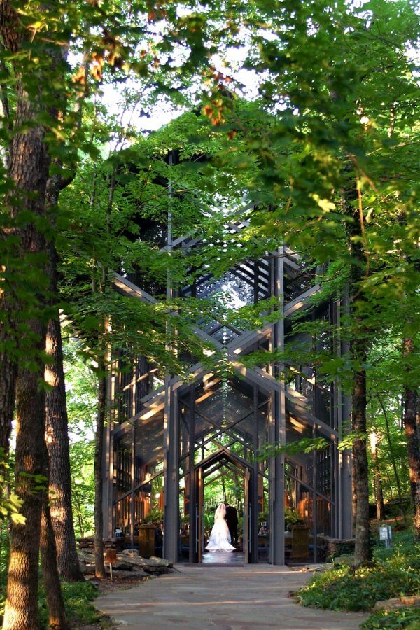 A bride and groom stand in one of the enchanting glass chapels of Arkansas, surrounded by lush green trees.