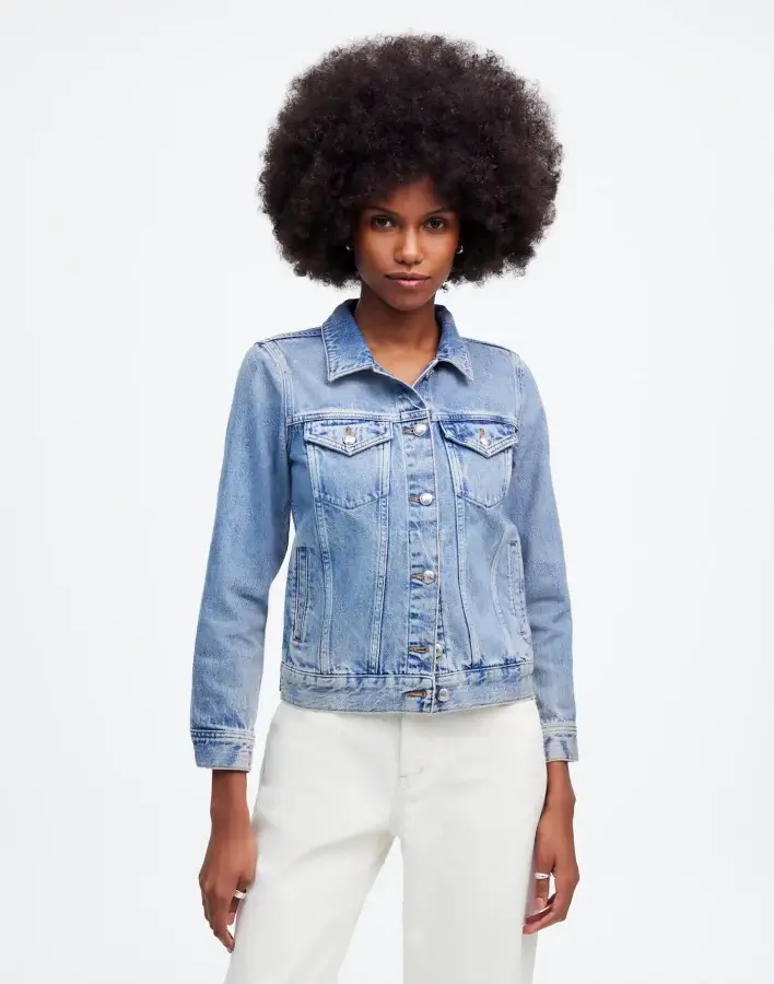 A person with an afro hairstyle embodies a modern spin on classic pieces, donning a light blue denim jacket and white pants, standing against a plain background. This look serves as a perfect addition to your spring wardrobe essentials.