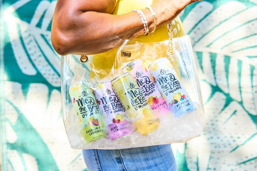 A person holds a transparent bag filled with ice and various canned flavored lemonades. The turquoise and white background features abstract leaf patterns.