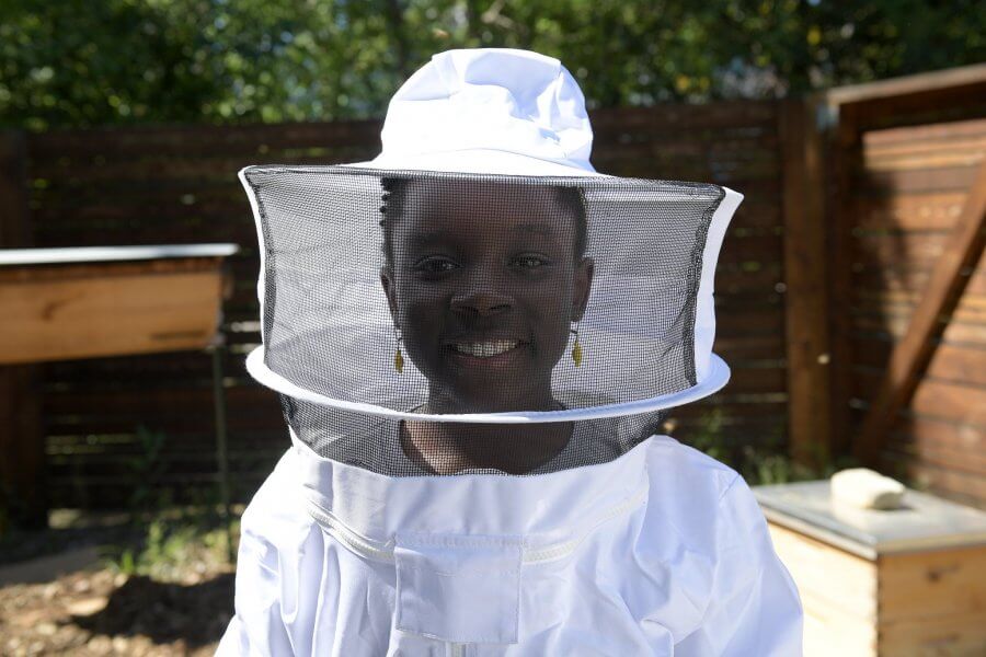 A person wearing a beekeeping suit with a mesh face cover stands outdoors near a wooden fence.