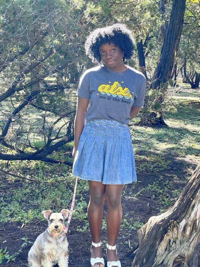 Mikaila Ulmer stands in a wooded area wearing a T-shirt and denim skirt, gently holding a small dog on a leash.