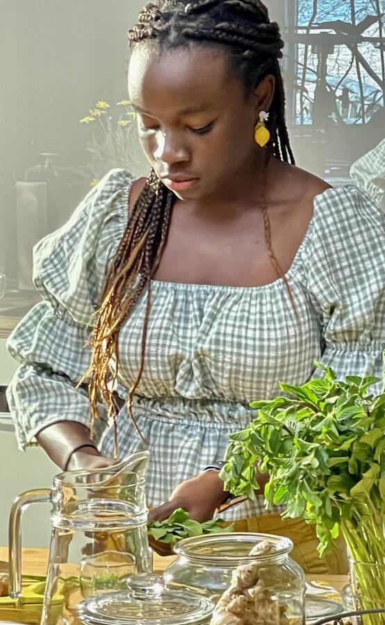 In the kitchen, surrounded by fresh herbs and a pitcher, a woman in a checkered dress prepares ingredients.