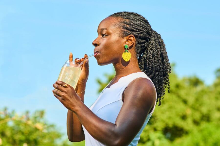 Mikaila Ulmer, in a white top, sips her Me & The Bees drink through a straw from a jar. Her braided hair is complemented by green and yellow earrings, set against a backdrop of blue sky and lush greenery.