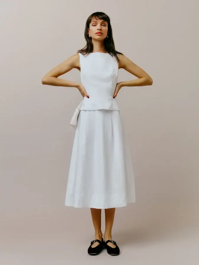 Person in a sleeveless white top and skirt, adding a modern spin with hands on hips against a neutral background, complemented by black shoes.