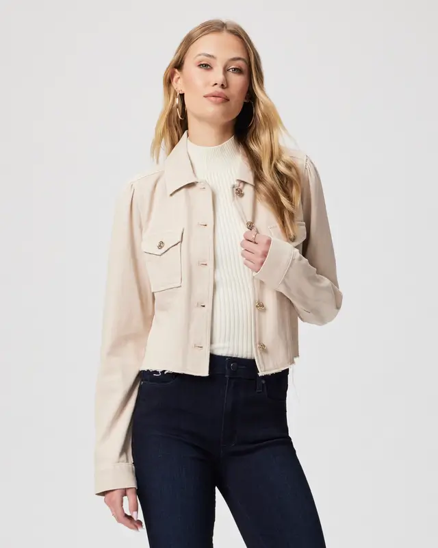 A woman, embodying spring wardrobe essentials, dons a cream-colored buttoned jacket over a white knit sweater and dark jeans. She stands against a plain white background, showcasing classic pieces with a modern spin that define timeless elegance.