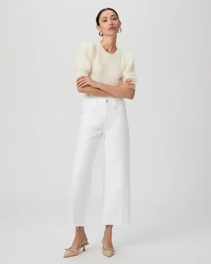 Person wearing a cream fluffy sweater, white wide-leg jeans, and beige heels, showcasing a modern spin on classic pieces. They're posing against a plain light background, exemplifying spring wardrobe essentials.