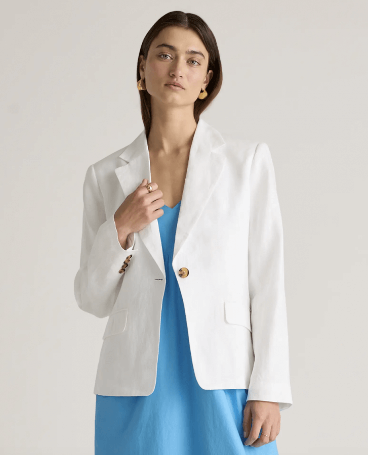 A person wearing a white blazer over a blue dress poses against a plain background, blending classic pieces with a modern spin.