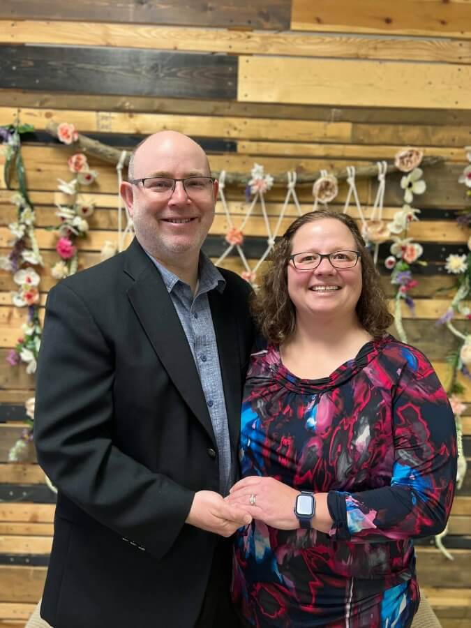 A man and a woman smiling while holding hands in front of a wooden wall adorned with flowers.