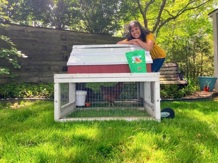 A person smiles while leaning on a small, wheeled chicken coop from Rent the Chicken. With trees and a wooden fence in the background, it's the perfect snapshot of backyard farming bliss.