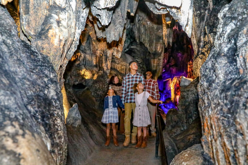 A family of four explores the captivating Alabama caves, admiring rock formations inside a dimly lit cavern with colorful lights in the background.
