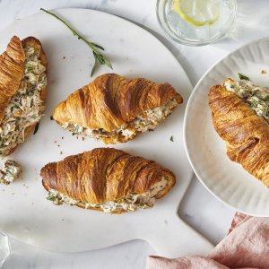 Three croissants filled with chicken salad on a white plate and board, with a pink napkin and glasses of lemon water nearby.