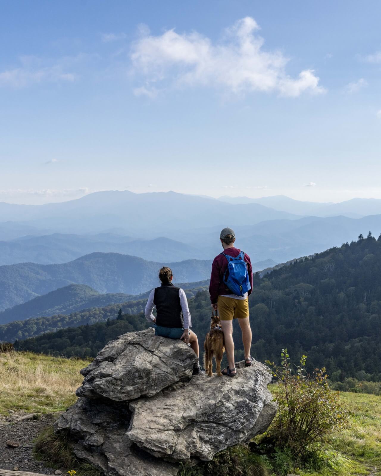Two people and their dog enjoy a family vacation in Tennessee, perched on a large rock on Roan Mountain overlooking the breathtaking mountainous landscape under a blue sky with clouds.