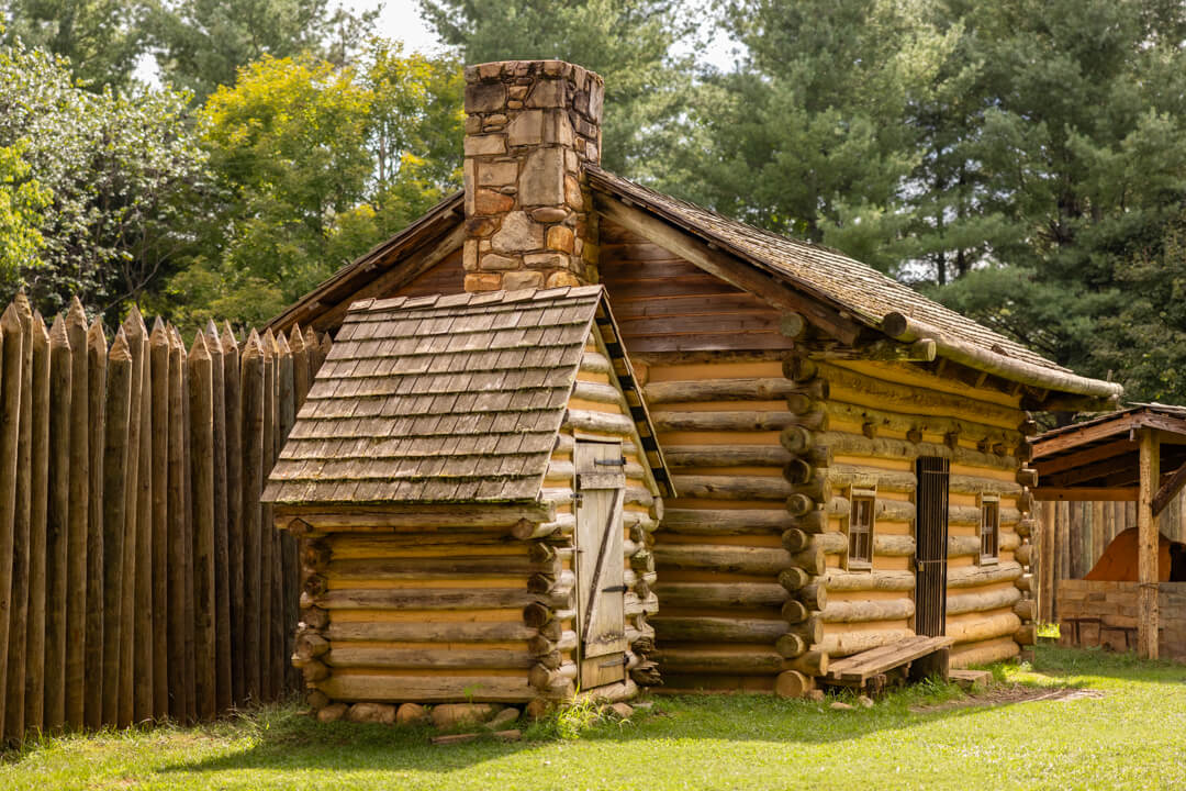 Nestled by a tall wooden fence in a wooded area, this log cabin Fort Watauga replica with a stone chimney is perfect for family vacations in Tennessee. It boasts a shingled roof and small windows, offering a cozy retreat amidst nature's beauty.