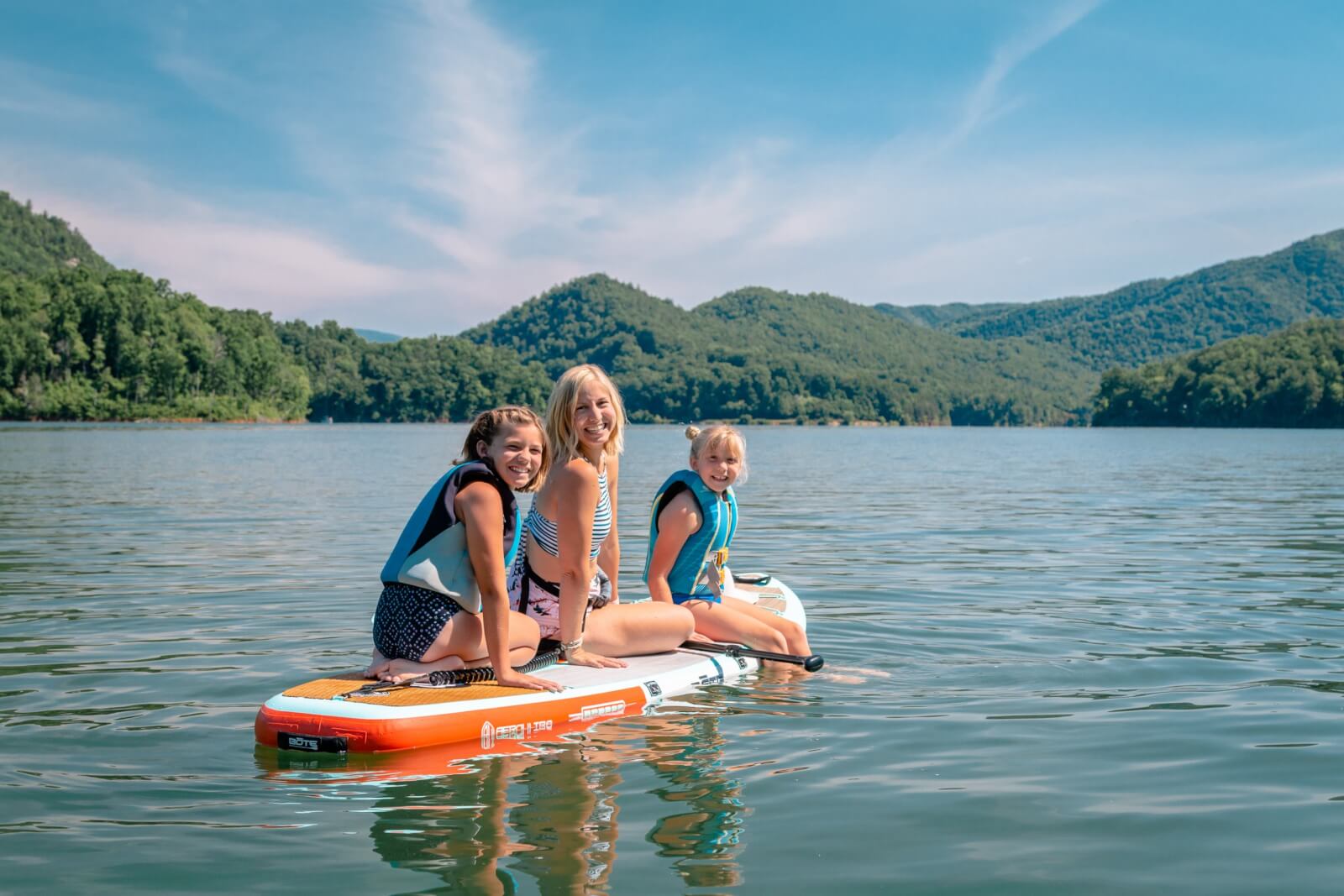 Three kids wearing life vests are sitting on a paddleboard in a calm lake, surrounded by the mountains of Tennessee under a clear blue sky, capturing the essence of family vacations.