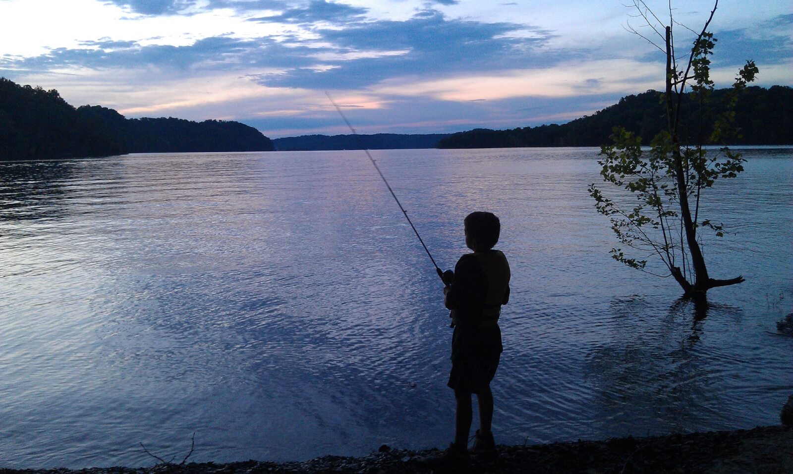A child is fishing at the edge of a calm Tennessee lake during dusk, capturing the serene essence of family vacations, with trees lining the distant shoreline and a partially cloudy sky above.