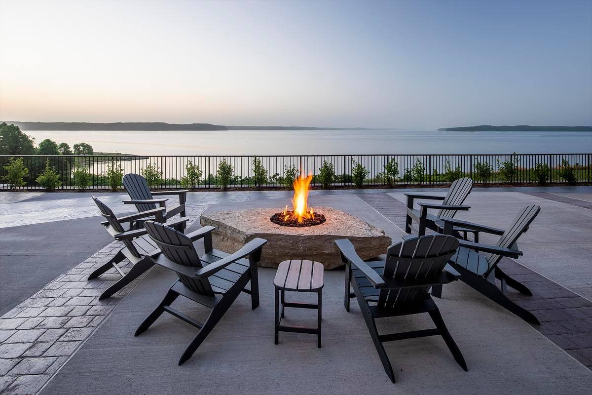 A circle of Adirondack chairs surrounds a lit stone fire pit on a patio, perfect for family vacations in Tennessee, overlooking a calm body of water. A railing frames the view of distant land under a clear sky.