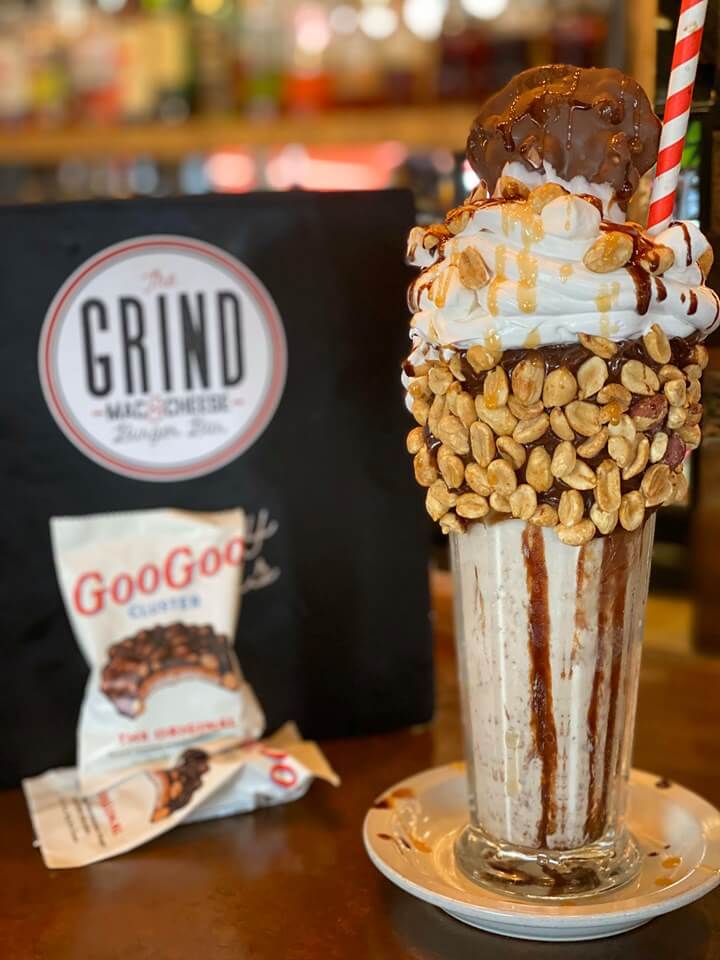 A tall milkshake with whipped cream, peanuts, and chocolate drizzle sits enticingly, topped with a chocolate treat and a sturdy straw. Beside it is a Goo Goo Cluster package. 