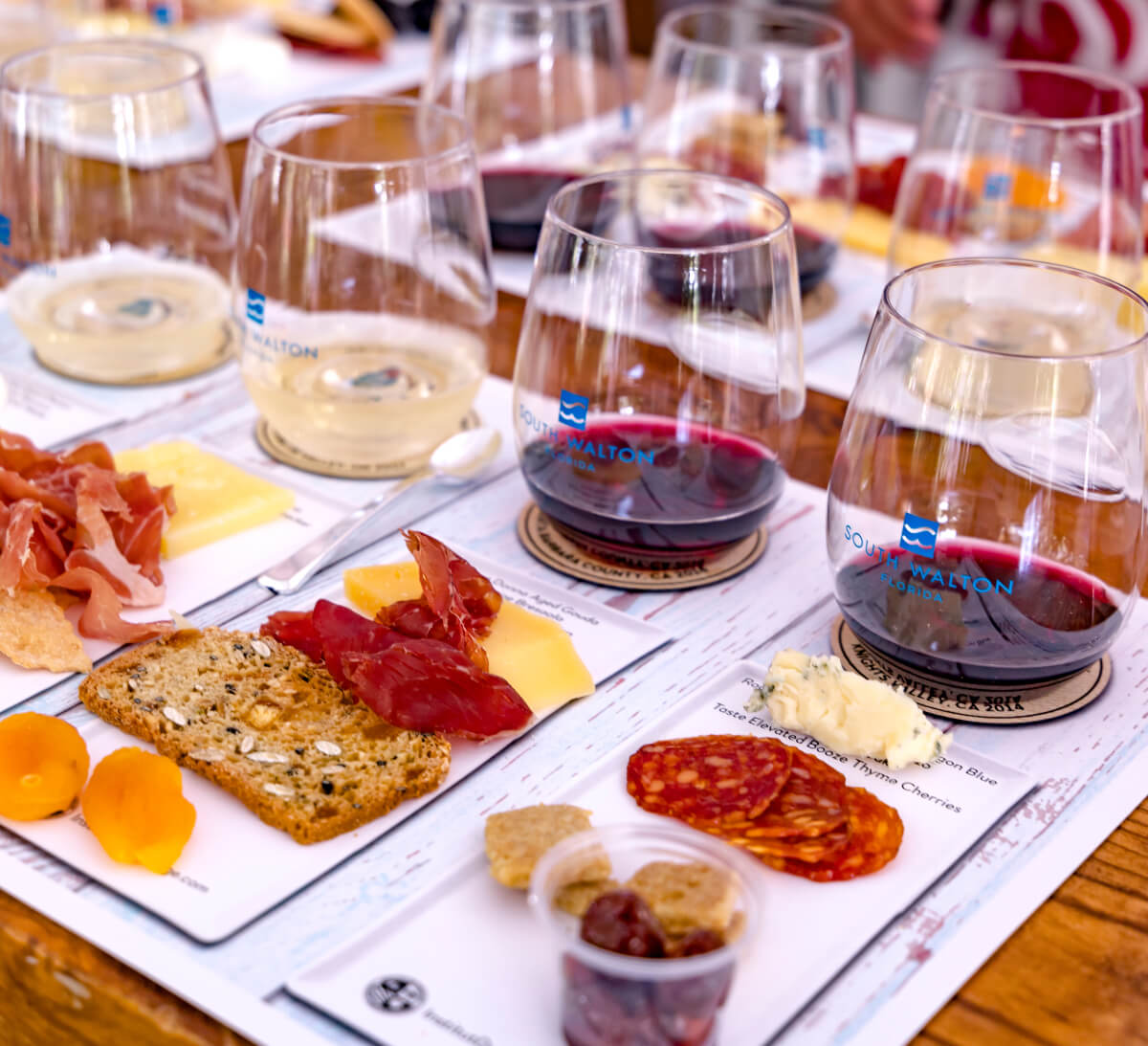 A wine and cheese tasting setup reminiscent of food and wine festivals, featuring glasses of red and white wine paired with assortments of cheese, cured meats, crackers, and dried apricots elegantly arranged on tasting mats.