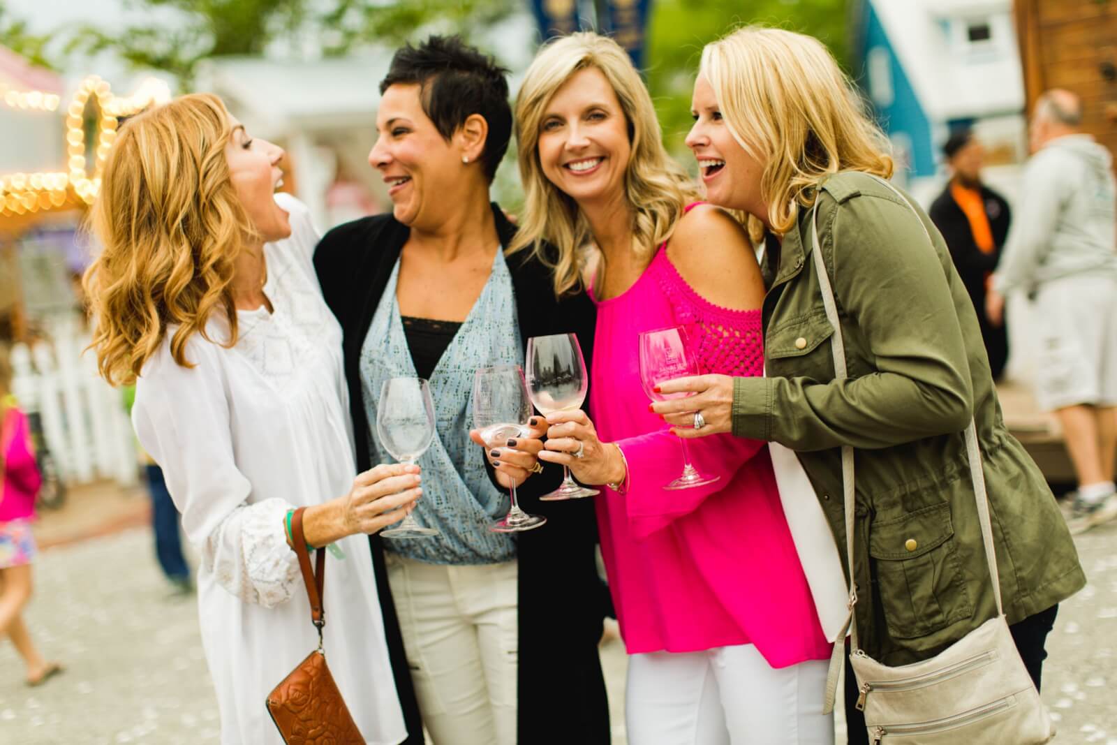 Four women are smiling and holding wine glasses at an outdoor food and wine festival.