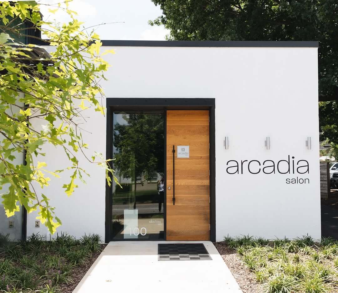The entrance of Arcadia Salon features a charming wooden door and glass side panel, embraced by lush greenery under Avondale's clear sky.
