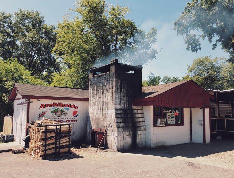 Small white building with a red roof and a tall chimney emitting smoke with the sign "Archibald's BBQ." A stack of firewood is piled nearby under sunny skies with trees in the background. 